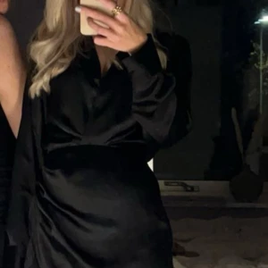 Svart satin klänning - Elegant svart klänning i satin med långärmad design och omlott-detalj. Perfekt för en stilren och sofistikerad look.