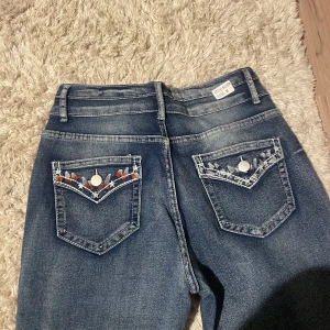 Blå bootcut jeans med broderade fickor - Snygga blå bootcut jeans från William Rast med unika broderade detaljer på bakfickorna. Jeansen har en klassisk femficksdesign och en bekväm passform. Perfekta för en avslappnad stil med en touch av personlighet.