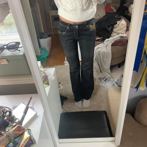 Gråa jeans med broderade detaljer - Snygga gråa jeans med unika broderade detaljer i gult och brunt på bakfickorna. Byxorna har en bootcut-stil och låg midja, vilket ger en avslappnad och trendig look. Perfekta för att ge din outfit en cool touch. Jag är 175cm.