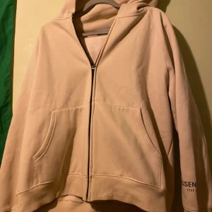 Beige hoodie från Essentials - Säljer en beige hoodie från Essentials med dragkedja och stor ficka framtill. Den har en luva och texten 'ESSENTIALS' tryckt på ryggen. Perfekt för en avslappnad stil.