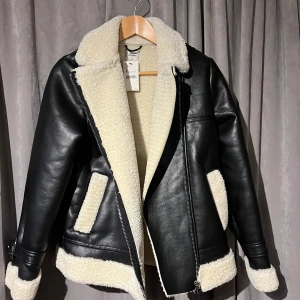 Svart skinnjacka/PU med teddyfoder XS. HELT NY - Snygg svart skinnjacka från Pimkie med mjukt vitt foder. Jackan har dragkedja och bälte vid ärmsluten. Perfekt för kyligare dagar! Helt ny!