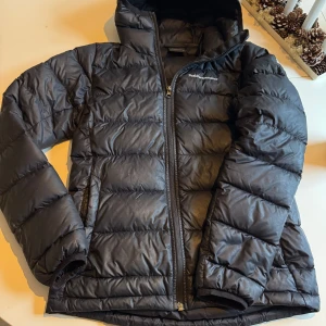 Svart dunjacka från Peak Performance - Snygg och varm svart dunjacka från Peak Performance med dragkedja och huva. Perfekt för små kyliga dagar med sin quiltade design och långa ärmar. Jackan har en stilren look och är idealisk för både stad och natur. Finns 3 små lagningar, inget som syns om man inte synar på nära håll.