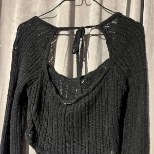 Svart stickad tröja med öppen rygg S - Snygg svart stickad tröja med långärmad design och öppen rygg. Tröjan har en knytning i nacken och ett ribbat mönster som ger en trendig look. Perfekt för lager-på-lager-stil eller som statement-plagg.