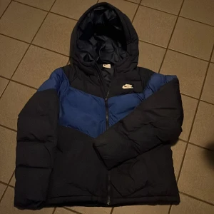 Blå dunjacka från Nike - Säljer en blå och svart dunjacka från Nike med huva och dragkedja. Jackan har en sportig look och är perfekt för kyligare väder. Den har en klassisk Nike-logga på bröstet och är designad för att hålla dig varm och bekväm. Fläck på höger framsida