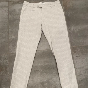 Randiga kostymbyxor från Les Deux - Snygga grå kostymbyxor från Les Deux med vita ränder. Byxorna har en slim passform med knappar och dragkedja i midjan. Perfekta för en stilren look.