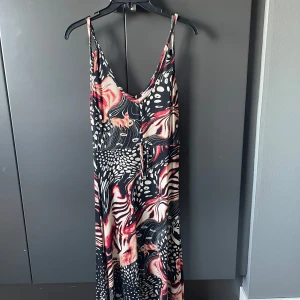 Mönstrad långklänning med knytband - Snygg långklänning med ett unikt mönster i svart, rött och beige. Klänningen har smala axelband och ett knytband framtill för extra detalj. Perfekt för en somrig look!