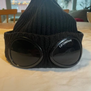 Svart  C.P. Company mössa, youth - Snygg svart ribbad mössa från C.P. Company med inbyggda glasögon på framsidan. Perfekt för att ge en unik touch till din outfit. Tillverkad i ull för extra värme och komfort.