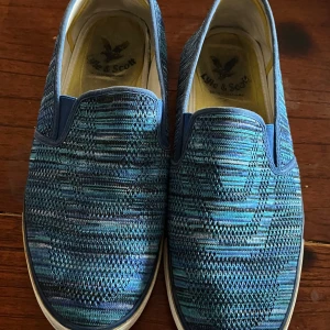 Blå slip-on skor från Lyle & Scott - Snygga blå slip-on skor från Lyle & Scott med ett stickat mönster. Skorna har en gul logotyp på hälen och en bekväm passform. Perfekta för en avslappnad stil.