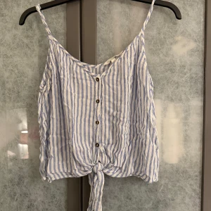 Randig topp från H&M - Säljer en somrig randig topp från H&M i blått och vitt. Toppen har knappar framtill och en knytning vid nederkanten. Perfekt för varma dagar!