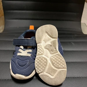 Mörkblå sneakers med kardborreband - Snygga mörkblå sneakers med vita snören och praktiskt kardborreband. Skorna har en sportig design med orange detaljer på hälen och en bekväm sula.