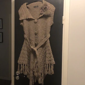 Grå stickad kofta/klänning  - Snygg grå stickad kofta/klänning från Seven Star med knappar och fransar. Den har en unik design med en stor krage och dekorativa blommor på axeln. Perfekt för att lägga till en bohemisk touch till din outfit. 35% ull. Passar både medium och large.