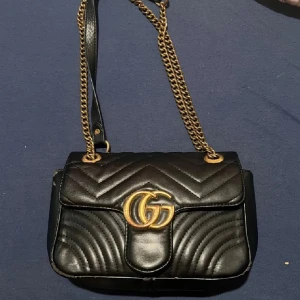 Svart quiltad axelväska med guldkedja - Snygg svart axelväska i skinn med quiltat mönster och guldfärgad kedja. Väskan har en elegant design med ett hjärtmönster på baksidan och en framträdande logotyp i guld på framsidan. Perfekt för att ge din outfit en lyxig touch. den är mycket användt