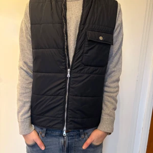 East West quiltad väst - Snygg svart quiltad väst med dragkedja och en praktisk ficka med knapp på bröstet. Perfekt för lager-på-lager under kyligare dagar. Enkel och stilren design som passar till det mesta. Modellen är 185 cm och väger 73 kg.