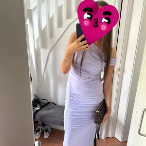 Ljuslila långklänning - Elegant ljuslila långklänning med offshoulder-design och smickrande passform. Klänningen har en stilren och modern look, perfekt för speciella tillfällen. Andvänd under en kväll