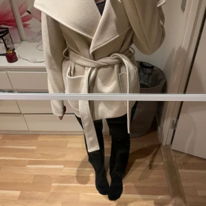 Beige kappa från Vero Moda - Säljer en stilren beige kappa från Vero Moda. Den har en elegant omlott-design med ett knytbälte i midjan och långa ärmar. Perfekt för att hålla stilen under kyligare dagar.