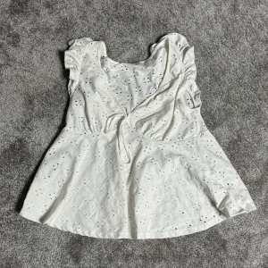 Vit blus med spetsdetaljer - Charmig vit blus med spetsdetaljer och volanger. Blusen har en v-ringad design med knytning framtill och är ärmlös. Perfekt för en somrig look.