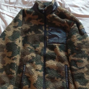 Camouflage teddyjacka från Saligia - Säljer en mysig teddyjacka i camouflage från Saligia. Jackan har en dragkedja framtill och en praktisk bröstficka. Perfekt för kyliga dagar med sin mjuka och varma känsla.