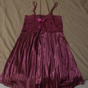 Vinröd klänning med spetsdetaljer - Charmig vinröd klänning med spetsdetaljer och satinliknande material. Den har smala axelband och en söt rosett framtill. Perfekt för en elegant look.