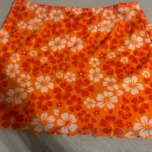 Orange blommig kjol från Shein - Säljer en färgglad orange kjol från Shein med ett härligt blommönster i vitt och rött. Kjolen är kort och perfekt för sommarens varma dagar. Den har en elastisk midja för en bekväm passform.