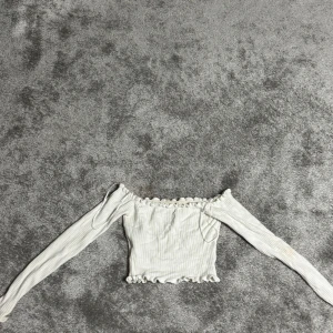 Vit ribbad offshoulder topp - Säljer en söt vit ribbad offshoulder topp med långa ärmar och volangdetaljer vid hals och nederkant. Perfekt för en stilren och trendig look. Passar bra till både jeans och kjol.