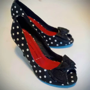 Svarta prickiga pumps med rosett - Charmiga svarta pumps med vita prickar och en söt rosett framtill. Skorna har en klack och en öppen tå, vilket ger en retro känsla. Insidan är klarröd, vilket ger en fin kontrast till det prickiga mönstret.