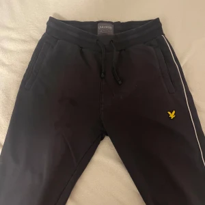Svarta mjukisbyxor från Lyle & Scott - Snygga svarta mjukisbyxor från Lyle & Scott med vit rand längs sidan och det klassiska gula logotypen. Byxorna har snörning i midjan för justerbar passform.