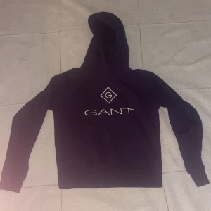 Svart hoodie från GANT - Snygg svart hoodie från GANT med vit logga på bröstet. Tröjan är i 176 cm för  16 år gamla.Tröjan har en fika på bröstet  och en bekväm huva. Perfekt för en avslappnad stil.