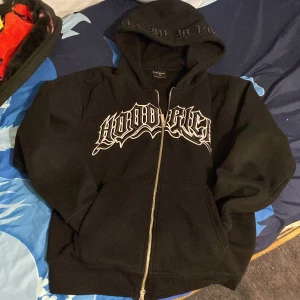 Svart hoodie från Hoodrich - Säljer en svart hoodie från Hoodrich med dragkedja och stor logga på framsidan. Huvan har textdetaljer och tröjan har en klassisk passform med fickor framtill. Perfekt för en avslappnad stil.
