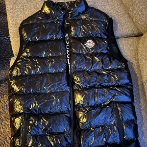 Svart dunväst från Moncler - Snygg svart dunväst från Moncler med glansigt material och dragkedja framtill. Västen har en hög krage och Moncler-logotypen broderad på bröstet. Perfekt för kyligare dagar,,, säljer den väldigt billigt .