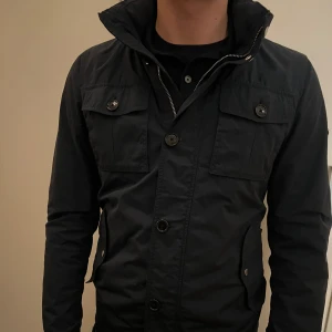 Svart jacka med dragkedja och knappar - Snygg svart jacka med både dragkedja och knappar framtill. Jackan har långa ärmar och praktiska fickor med knappar på bröstet. Perfekt för en stilren look.