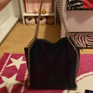 Svart axelväska från Tiamo - Snygg svart axelväska från Tiamo med kedjedetaljer längs kanterna. Väskan har en dragkedja på baksidan och är tillverkad i syntetmaterial. Lite gammal stockholm stil men den är fortfarande perfekt för små barn som börjar komma in i stilen