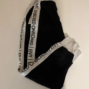 Svarta mjukisbyxor med vita detaljer från Envy - Säljer ett par svarta mjukisbyxor från Envy med vita detaljer och text längs sidorna. Byxorna har en bekväm passform och elastiska muddar vid bensluten. Perfekta för en avslappnad stil.