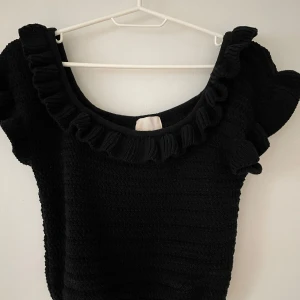 Svarta stickad topp med volangdetaljer - Säljer en svart stickad topp med volangdetaljer runt halsringningen. Toppen har en kortärmad design och är perfekt för en stilren look. Passar bra till både jeans och kjol.