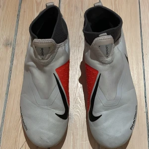 Nike Phantom Vision fotbollsskor - Säljer ett par ljusgråa Nike Phantom Vision fotbollsskor med röd träffyta. Skorna har strumpa och en gömd snörning under skalet så kallad ”Ghost lace” och är perfekta för fotbollsplanen. De har en slitstark multiground sula med dobbar för optimalt grepp.