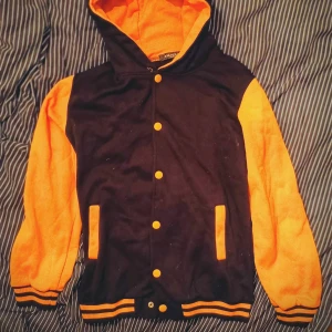Svart och orange/gul vintage huvjacka - Snygg svart och orange/gul huvjacka med knappar och huva. Jackan har långa ärmar och ribbade muddar i matchande färger. Perfekt för en sportig vintage  look. [Obs! Knappen längst ned går ej att stänga men det går bara att byta ut mot en ny.]