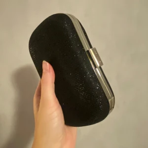 Svart glittrig kuvertväska från Karen Millen - Snygg svart kuvertväska från Karen Millen med glittrig yta och silverfärgad ram. Perfekt för festliga tillfällen! Väskan har en smidig stängning och är i mycket bra skick. En stilren accessoar som passar till många outfits. Utan axelband. 