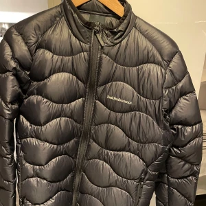 Svart dunjacka från Peak Performance - Säljer en snygg svart dunjacka från Peak Performance. Den har en quiltad design med dragkedja framtill och är perfekt för kyliga dagar. Jackan är i mycket bra skick och passar både till vardags och utomhusaktiviteter. Perfekt för höst och vinter!