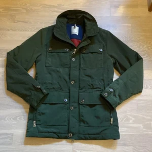 J.lindberg jacka - Säljer en snygg grön jacka i mycket bra skick. Den har långa ärmar och är perfekt för höst, vinter och vår. Jackan har både knappar och dragkedja framtill, vilket ger extra stil och funktionalitet. Den är gjord av ett slitstarkt material och har flera praktiska fickor.