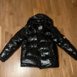 Säljer en snygg svart pufferjacka från Moncler i nytt skick. Jackan är perfekt för vintern med sitt blanka polyester-material och långa ärmar. Den har en praktisk huva och dragkedja framtill. Passar både till vardags och festliga tillfällen. Storleken är Large och passar perfekt för mig som är 183cm lång! 