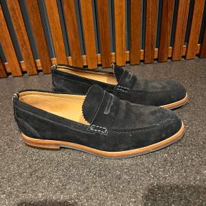 Snygga marinblå loafers i mocka från Hudson London. De har en klassisk design med en stilren söm och en bekväm innersula. Perfekta för både vardag och festliga tillfällen. Sulan är i ljust material som ger en fin kontrast till det mörka mockaskinnet.