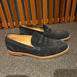Marinblå mockaloafers från Hudson London - Snygga marinblå loafers i mocka från Hudson London. De har en klassisk design med en stilren söm och en bekväm innersula. Perfekta för både vardag och festliga tillfällen. Sulan är i ljust material som ger en fin kontrast till det mörka mockaskinnet.