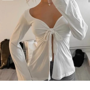 Vit långärmad topp med knyt - Säljer en superfin vit långärmad topp med knyt framtill. Den har en snygg v-ringad design och är perfekt för både vardag och fest. Materialet känns mjukt och bekvämt, och den har en lite lös passform som gör den extra skön att bära. Passar perfekt till jeans eller kjol!