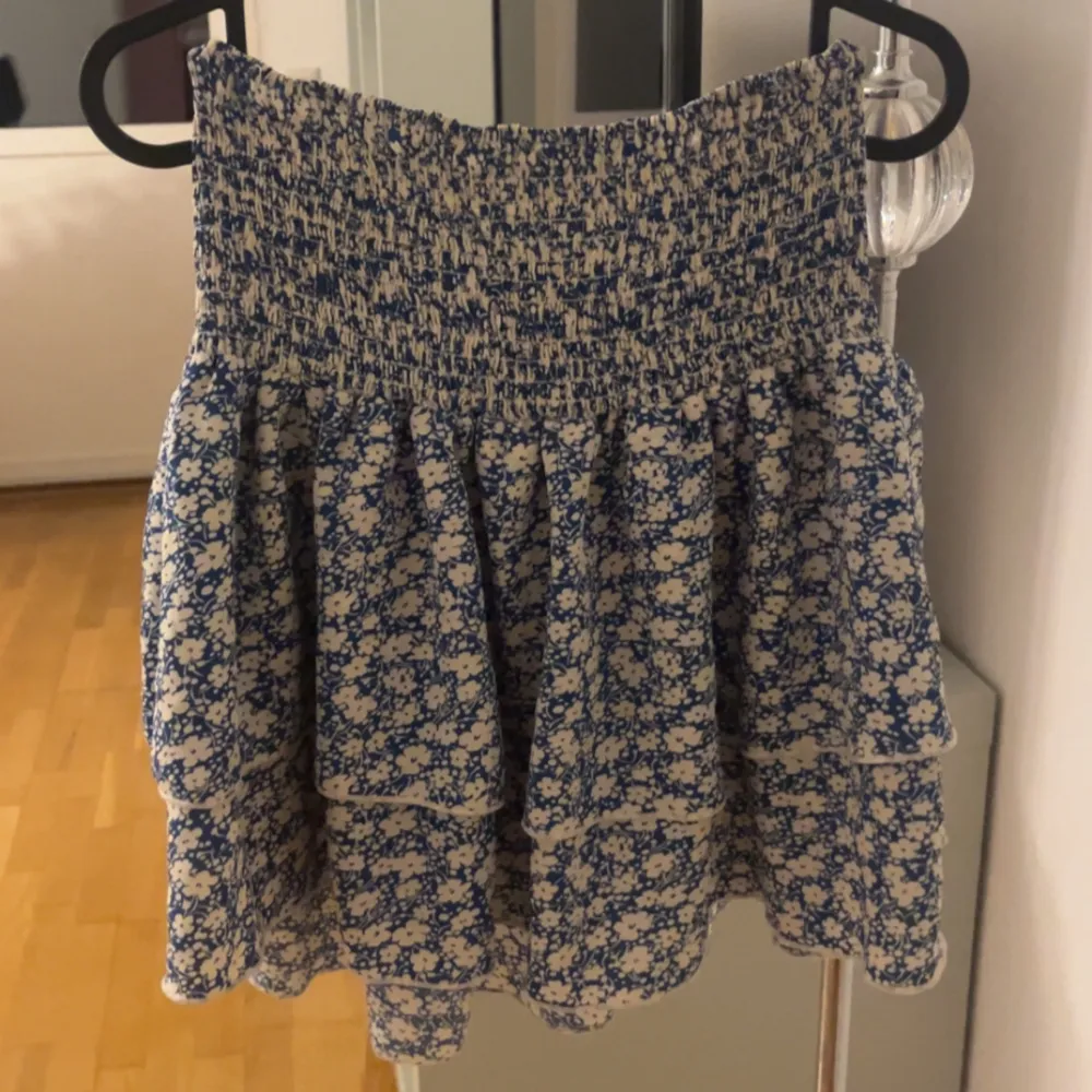 Söt blommig kortkjol i blått och vitt med volanger. Perfekt för sommardagar eller en avslappnad vårlook. Den har en elastisk midja för en bekväm passform.. Kjolar.