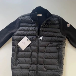 Moncler Cardigan - Säljer en snygg svart Moncler cardigan i nytt skick (äkta). Jackan har långa ärmar och en dragkedja framtill. Perfekt för höst och vinter med sitt quiltade mönster och stilrena design. Moncler-loggan är broderad på ärmen (NFC- Scan). Passar både till vardags och lite finare tillfällen.