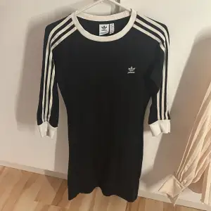 Snygg svart klänning från Adidas med klassiska vita ränder på ärmarna. Den har en sportig look med ribbade muddar och rund halsringning. Perfekt för en avslappnad stil eller sportiga tillfällen. Klänningen är i bra skick och passar perfekt för höst och vår.