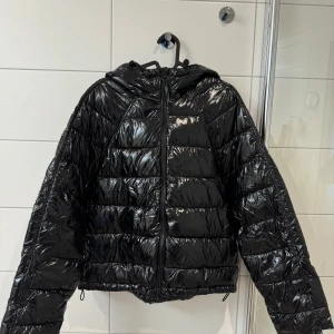 Svart glansig pufferjacka från Zara - Säljer en svart glansig pufferjacka från Zara i storlek S. Jackan har en dragkedja framtill och en mysig huva. Perfekt för kyliga  höst- och vinterdagar. Har dragsko i midjan och knappar i ärmsluten. Den är i mycket bra skick och redo för nya äventyr! 🖤