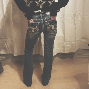 Jeans med broderad ficka - Säljer ett par snygga jeans i bra skick med coola broderade detaljer på bakfickorna. De är blå med en lätt slitning och har en bootcut-stil. Perfekta för en avslappnad look med en touch av unik stil. fläck på knät. visas på bilden