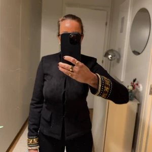 Svart stretch kavaj med broderade detaljer - Säljer en elegant svart jacka med unika broderade detaljer i guld och brunt på ärmsluten. Jackan har en figurnära passform och är perfekt för höst och vår. Den är långärmad och har en stilren design som passar både till vardags och fest. Perfekt för dig som vill sticka ut med stil!