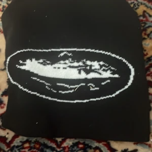 Svart mössa med broderad detalj - Säljer en svart mössa med en cool broderad detalj på framsidan. Perfekt för att hålla sig varm under höst och vinter. Mössan är i bra skick och passar de flesta huvudstorlekar. Enkelt och stilrent val för kalla dagar!