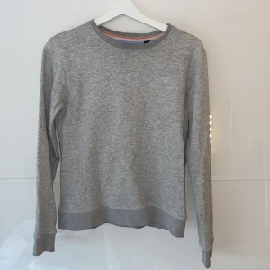 Grå tröja från GANT - Säljer en stilren grå tröja från GANT i storlek 170cm. Den är supermjuk och perfekt för både vardag och chill. Tröjan har långa ärmar och en klassisk rund halsringning. Diskret GANT-logga broderad på bröstet. Passar perfekt till jeans eller chinos!